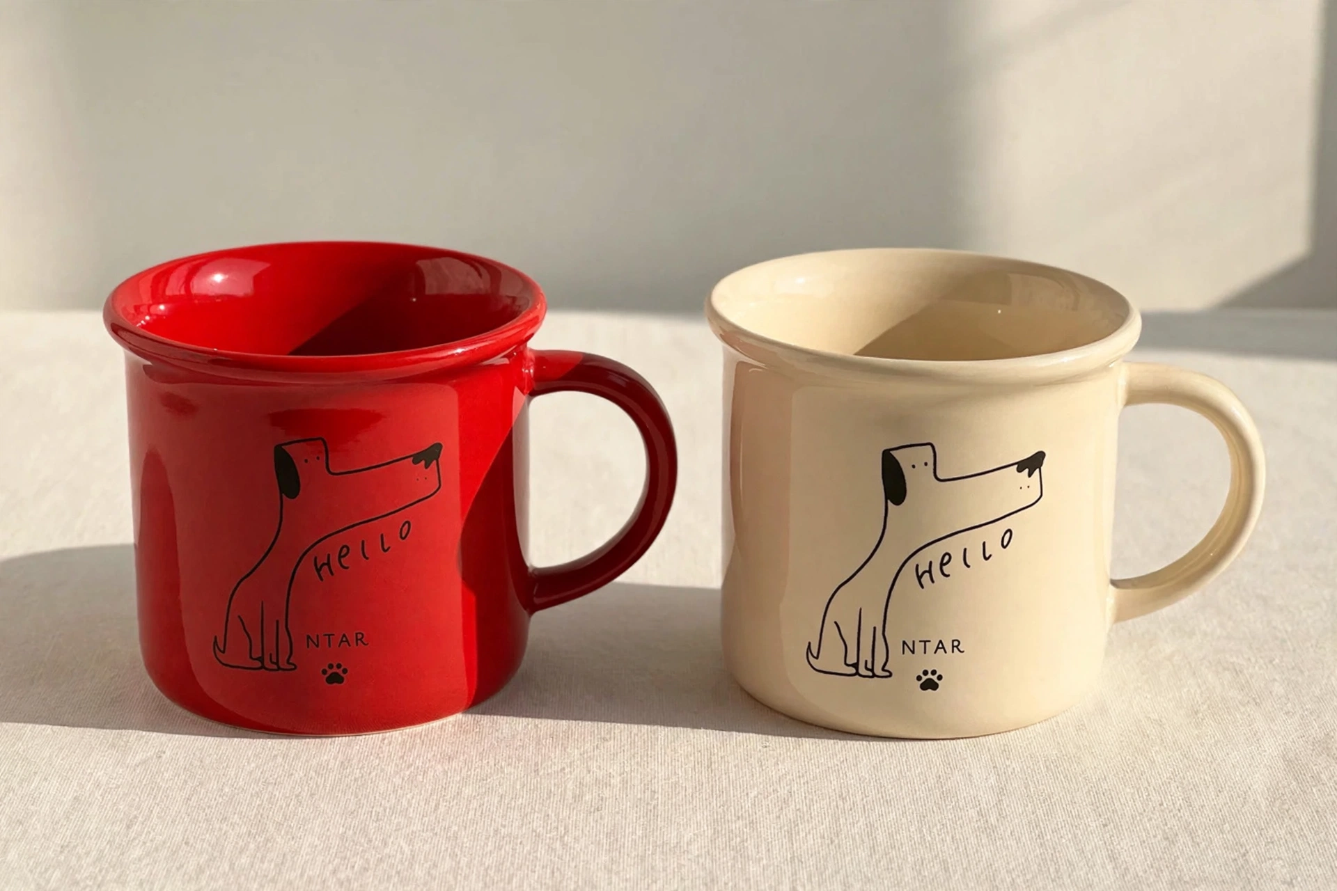 Pair of ceramic mugs, one wine-red and one cream, each with a line-art dog, the word Hello, and a small NTAR paw logo