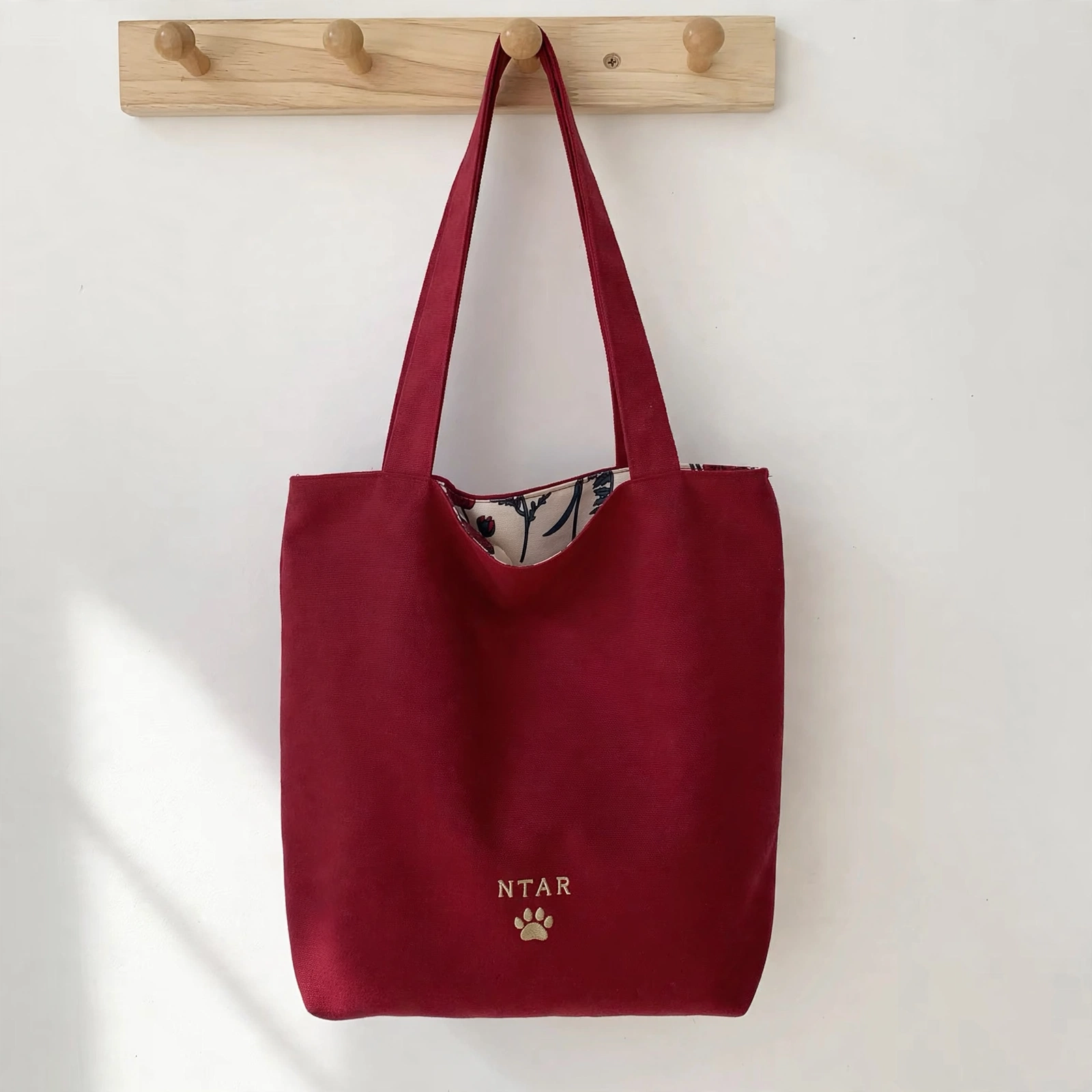 Wine-red canvas tote bag hanging on a wooden peg rail, solid wine-red side out with a small embroidered NTAR paw logo and the floral reverse peeking at the top
